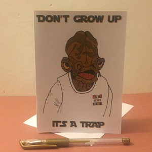 Puede incluir: Tarjeta de felicitación con un personaje de dibujos animados y el texto "DON'T GROW UP" y "IT'S A TRAP". Un bolígrafo dorado está delante de la tarjeta. La tarjeta está sobre una superficie blanca.