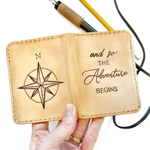 Puede incluir: Un diario de cuero marrón con un diseño de brújula y el texto "and so the adventure begins" en la portada.