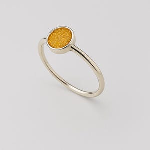 Könnte beinhalten: Ein minimalistischer Ring mit einem runden, strukturierten, goldgelben Stein, der in einem dünnen, silberfarbenen Band gefasst ist. Das Design des Rings ist schlicht und elegant, mit Fokus auf dem Kontrast zwischen dem hellen Stein und dem polierten Metall.