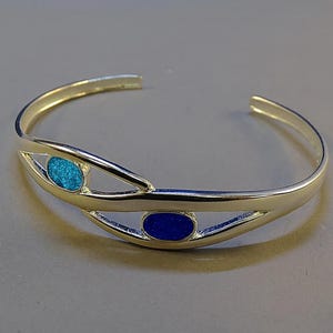 May include: A silver-toned cuff bracelet with an open design. The bracelet features two oval-shaped insets, one filled with turquoise-colored material and the other with a deep blue material. The bracelet is displayed on a gray surface.