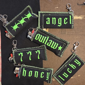 May include: Five black fabric lighter sleeves with neon green stitching and text. The sleeves feature the words angel, outlaw, 777, honey, and lucky in a gothic font, along with a barbed wire graphic. Each sleeve includes a silver metal lobster clasp.