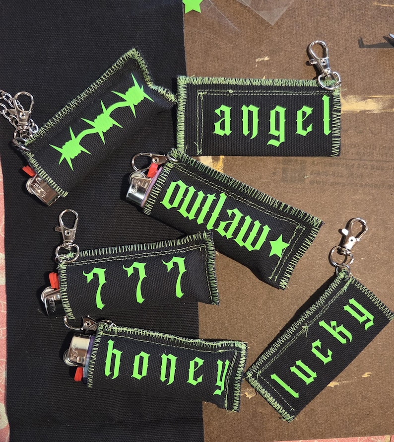 May include: Five black fabric lighter sleeves with neon green stitching and text. The sleeves feature the words angel, outlaw, 777, honey, and lucky in a gothic font, along with a barbed wire graphic. Each sleeve includes a silver metal lobster clasp.