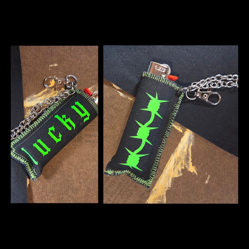 May include: A black fabric lighter sleeve featuring a bright green barbed wire graphic on one side and the word "lucky" in a gothic font on the other. The sleeve includes a silver-toned metal chain with a lobster clasp attachment.