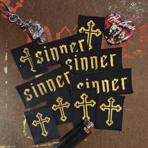 May include: A collection of black fabric patches featuring gold embroidered crosses and the word sinner in a gothic font. The set includes a silver skull keychain, a heart-shaped pin with The Hex Girls text, and a black Bic lighter.