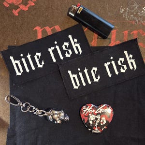 May include: Two rectangular black fabric patches with the white text bite risk, a black Bic lighter, a silver-toned skull keychain, and a heart-shaped pin featuring an illustration of The Hex Girls with the text Scooby-Doo.