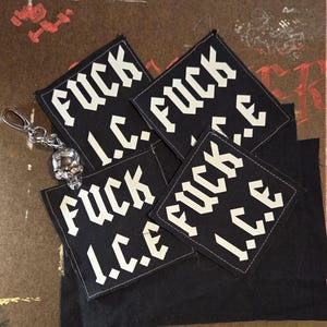 May include: Three square black fabric patches featuring the white text FUCK I.C.E. in a bold, angular font. A silver-toned metal skull keychain with a lobster clasp is attached to the top left patch, resting on a dark, textured fabric surface.