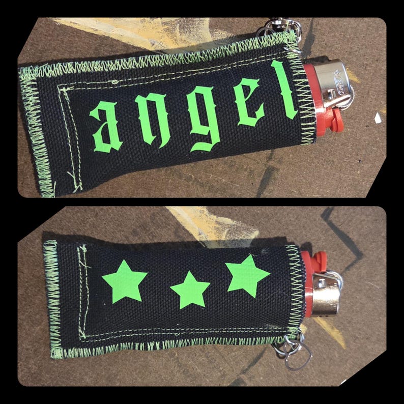 May include: A black fabric lighter sleeve with lime green stitching and the word angel in a gothic font on one side. The reverse side features three lime green star shapes. The sleeve is fitted over a red lighter with a silver metal top.