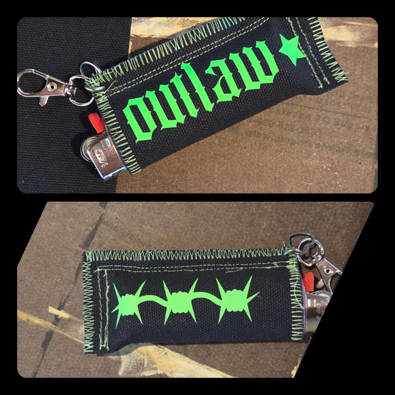 May include: A black fabric lighter sleeve featuring a neon green stitched border and a silver metal lobster clasp. One side displays the word outlaw in a gothic font with a star, while the other side features a neon green barbed wire graphic.