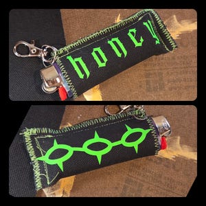 May include: A black fabric lighter sleeve featuring a silver metal lobster clasp. One side displays the word honey in bright green gothic-style lettering, while the other side features a graphic of three connected green spiked circular shapes.