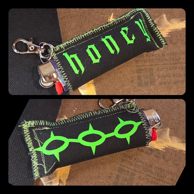 May include: A black fabric lighter sleeve featuring a silver metal lobster clasp. One side displays the word honey in bright green gothic-style lettering, while the other side features a graphic of three connected green spiked circular shapes.
