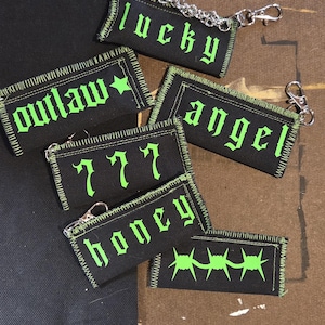 May include: A collection of rectangular black fabric keychains with neon green embroidered text and graphics. The designs include the words lucky, outlaw, angel, 777, and honey, plus a barbed wire graphic. Each piece features a silver metal chain.