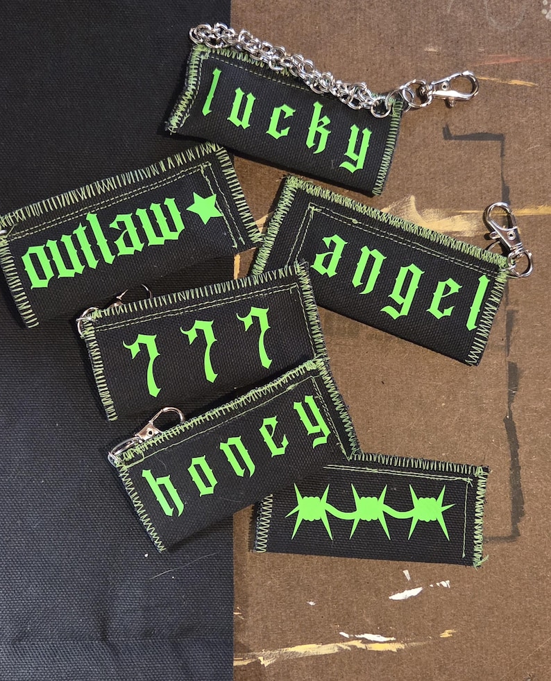 May include: A collection of rectangular black fabric keychains with neon green embroidered text and graphics. The designs include the words lucky, outlaw, angel, 777, and honey, plus a barbed wire graphic. Each piece features a silver metal chain.