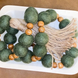 Puede incluir: Guirnalda de cuentas decorativa con bolas de fieltro verde oliva y cuentas de madera natural. La guirnalda está adornada con una borla de yute y se exhibe en una bandeja blanca. Ideal para la decoración del hogar.