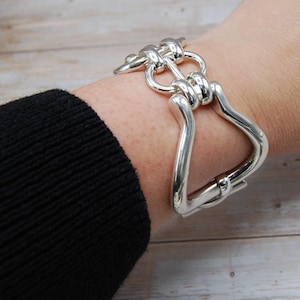 Op de afbeelding: Een zilverkleurige armband met een uniek ontwerp. De armband heeft een groot, gebogen, U-vormig element dat is verbonden met een reeks in elkaar grijpende, afgeronde schakels. De armband wordt om een pols gedragen.