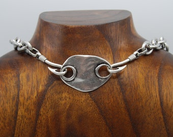 May include: A silver necklace featuring a large, flat, oval pendant with two small, round rings. The pendant has a textured surface and the chain is made of thick, linked metal.