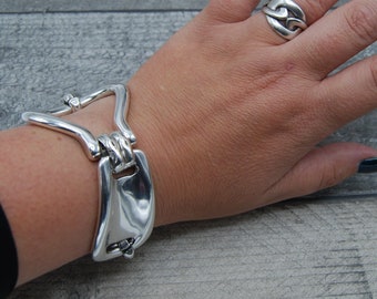 May include: A silver bracelet with a unique, open-link design. The bracelet is shown on a person's wrist, along with a silver ring with a similar design.