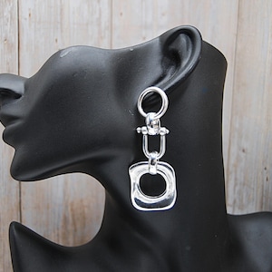 May include: Silver-coloured dangle earrings with a geometric design. The earrings feature a circular top, a U-shaped middle section, and a square bottom. The earrings are displayed on a black mannequin head against a light wood background.
