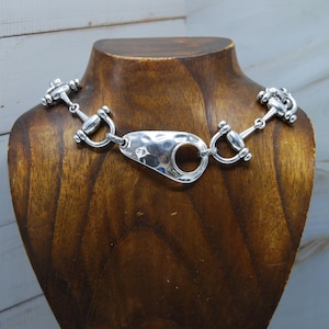 May include: Silver necklace with a horse bit design. The necklace has a hammered central piece with a large circular opening, connected to bit-shaped elements. The necklace is displayed on a wooden bust.