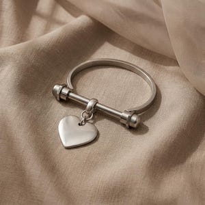 May include: A silver bracelet featuring a heart-shaped pendant. The bracelet has a hinged design with a bar and screw closure. The heart charm is suspended from a small ring. The bracelet is shown on a beige fabric.