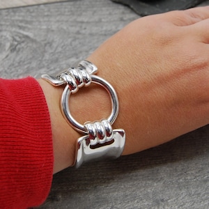May include: A silver bangle bracelet with a large, round, open circle design. The bracelet is made of thick, polished metal and has a modern, minimalist style.