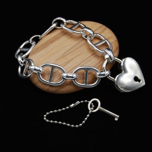 May include: A silver bracelet with a heart-shaped padlock and a matching key. The bracelet is made of a thick, chunky chain with a nautical-inspired design.