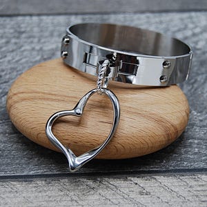 May include: A silver metal collar with a heart-shaped pendant. The collar is wide and has rivets along the edges. The heart pendant hangs from a small chain attached to the collar's centre. The items rest on a wooden, oval-shaped surface.