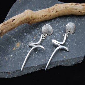 May include: A pair of silver-toned dangle earrings. Each earring features a hammered disc, a curved, abstract design, and a long, slender, curved bottom. The earrings are displayed on a dark grey stone surface with a piece of driftwood.