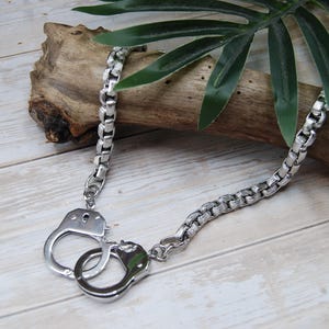 May include: A silver-colored necklace featuring a pair of handcuffs as a pendant. The necklace has a box chain design. The handcuffs are detailed with keyholes and a locking mechanism. The jewelry is displayed on a wooden surface with a green leaf.