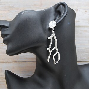May include: Silver earrings with a branch design. Each earring has a small, round stud and a long, textured, branch-shaped pendant. The earrings are shown on a black mannequin head against a light wood-grain background. A stylish accessory.