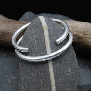 May include: Two silver bangle bracelets, one slightly larger than the other, are stacked on top of each other. The bracelets are resting on a grey stone with a white stripe running down the centre.