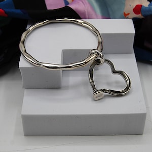 May include: A silver bracelet with a textured, wavy design. A heart-shaped charm hangs from the bracelet. The bracelet is displayed on a white surface, with a colourful fabric in the background. The bracelet is a piece of jewellery.