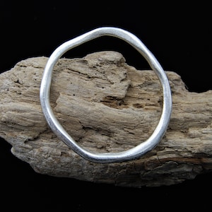 May include: A silver bangle bracelet with an irregular, organic shape. The bracelet is resting on a piece of driftwood.