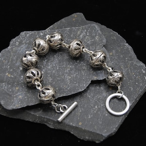 May include: Silver bracelet featuring ornate, hollow, spherical beads linked together. The bracelet has a toggle clasp and is displayed on a dark gray, textured stone against a black background. The beads have intricate cut-out designs.