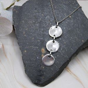 May include: A silver necklace featuring three circular pendants suspended from a delicate chain. The pendants are evenly spaced and have a slightly hammered texture. The necklace is displayed on a dark gray stone surface.