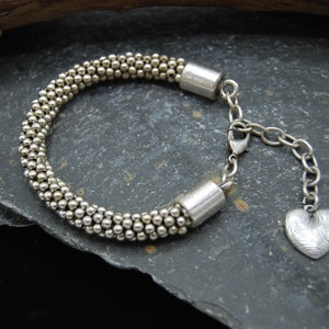 May include: A silver bracelet with a heart charm. The bracelet is made of small silver beads and has a clasp closure. The heart charm is textured and has a slightly hammered finish.