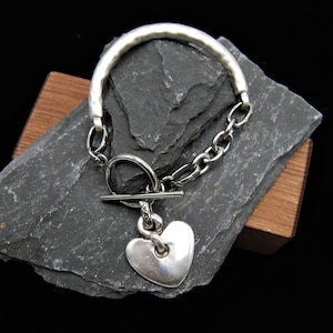 Pode incluir: Uma pulseira de prata com um fecho de coração e um fecho de correr. A pulseira é feita de uma banda grossa de prata texturizada e um elo de corrente.