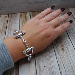 Puede incluir: Un brazalete plateado con un diseño de bocado de caballo. El brazalete de metal presenta una serie de eslabones conectados en forma de bocado. El brazalete se lleva en la muñeca, mostrando su estilo ecuestre.