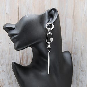 Könnte beinhalten: Ein silberner Ohrring mit einem einzigartigen Design. Der Ohrring hat einen kreisförmigen oberen Teil, ein rechteckiges Glied mit einem dekorativen Element und einen langen, spitz zulaufenden Anhänger. Der Ohrring wird auf einem schwarzen Schaufensterkopf vor hellem Holz gezeigt.