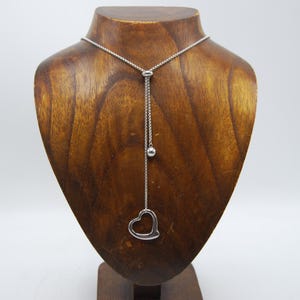 Puede incluir: Collar plateado con un colgante en forma de corazón, exhibido en un busto de madera. El colgante es un corazón abierto. El collar tiene una cadena fina y una pequeña cuenta esférica. El busto es de madera.