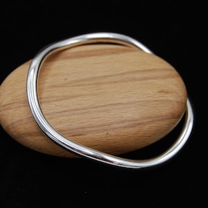 Könnte beinhalten: Ein silbernes Armreif-Armband mit einer glatten, abgerundeten Oberfläche. Das Armband wird auf einer Holzoberfläche präsentiert.