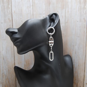 May include: Silver-toned dangle earrings with a geometric design. Each earring features a circular stud, connected to a series of elongated, oval, and rectangular shapes. The earrings are displayed on a black mannequin head against a light wood background.