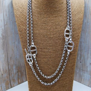 Puede incluir: Un collar plateado con un diseño de doble cadena. El collar presenta una serie de eslabones circulares y elementos decorativos. El collar se exhibe sobre un maniquí marrón, sobre un fondo de madera clara.
