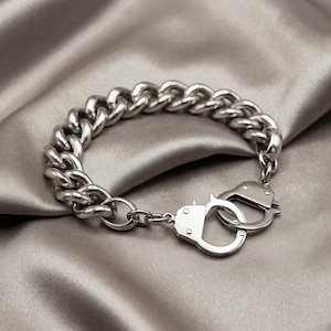 Könnte beinhalten: Ein silberfarbenes Armband mit einem klobigen Kettendesign und einem Handschellenverschluss. Das Armband aus glänzendem Metall liegt auf einem Satin-Stoff.