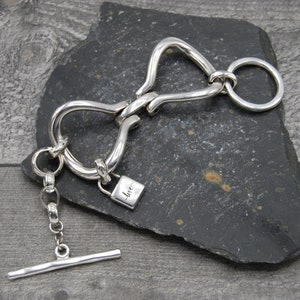 May include: Silver bracelet featuring a padlock charm inscribed with the word "love" and a toggle clasp. The bracelet is crafted from thick, twisted metal.