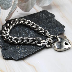 Könnte beinhalten: Ein silberfarbenes Kettenarmband mit herzförmigem Vorhängeschloss-Verschluss. Das Armband hat ein klobiges Panzerketten-Design. Das herzförmige Vorhängeschloss hat ein Schlüsselloch. Das Armband wird auf einem dunkelgrauen Stein präsentiert.