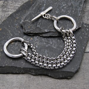 May include: A silver chain bracelet with multiple chain links and a toggle clasp. The bracelet has two large, round, silver rings.