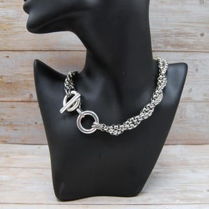 May include: A silver necklace with a toggle clasp, presented on a black mannequin. The necklace has a thick, braided chain design. The toggle clasp is a circular ring with a bar closure.