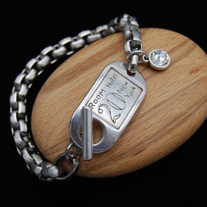 May include: A silver bracelet with a silver charm that says "Room Hotel 20 New York". The charm has a small silver loop and a clear rhinestone attached to it.