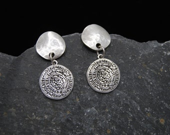 May include: A pair of silver-toned earrings. Each earring features a hammered, disc-shaped top and a circular pendant with an engraved pattern. The earrings are displayed on a dark gray, textured surface.