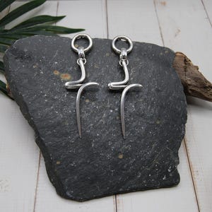 Puede incluir: Un par de pendientes plateados con un diseño abstracto único. Cada pendiente presenta una parte superior circular conectada a una forma curva y alargada. Los pendientes se exhiben sobre una piedra gris oscura contra un fondo de madera blanca.